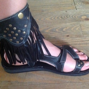 Puede incluir: Sandalias de cuero negro con detalle de flecos y una correa de tobillo con tachuelas doradas.