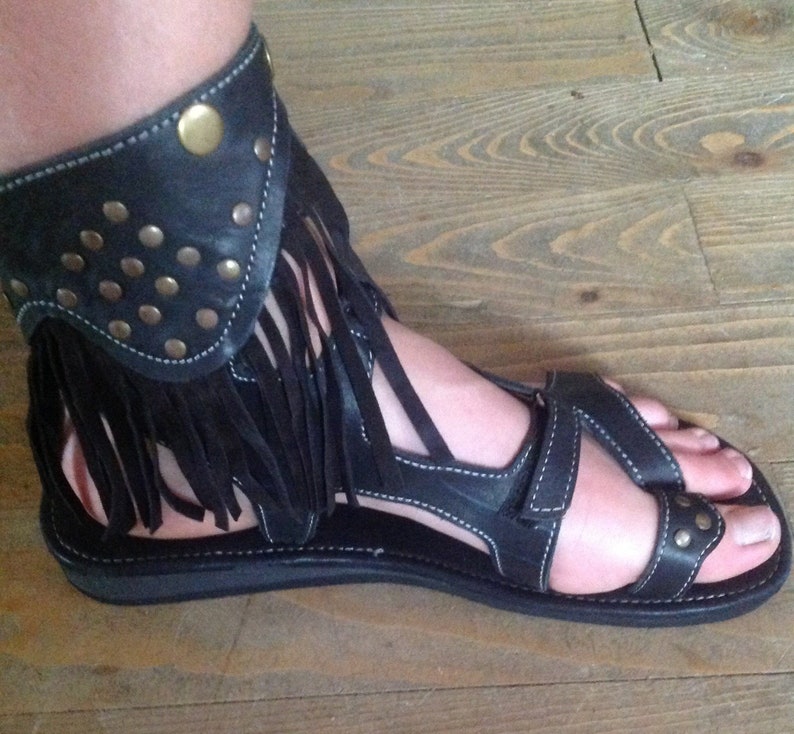 Puede incluir: Sandalias de cuero negro con detalle de flecos y una correa de tobillo con tachuelas doradas.