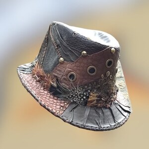 May include: A brown leather hat with a wide brim and a variety of textures and embellishments, including feathers, studs, and eyelets.