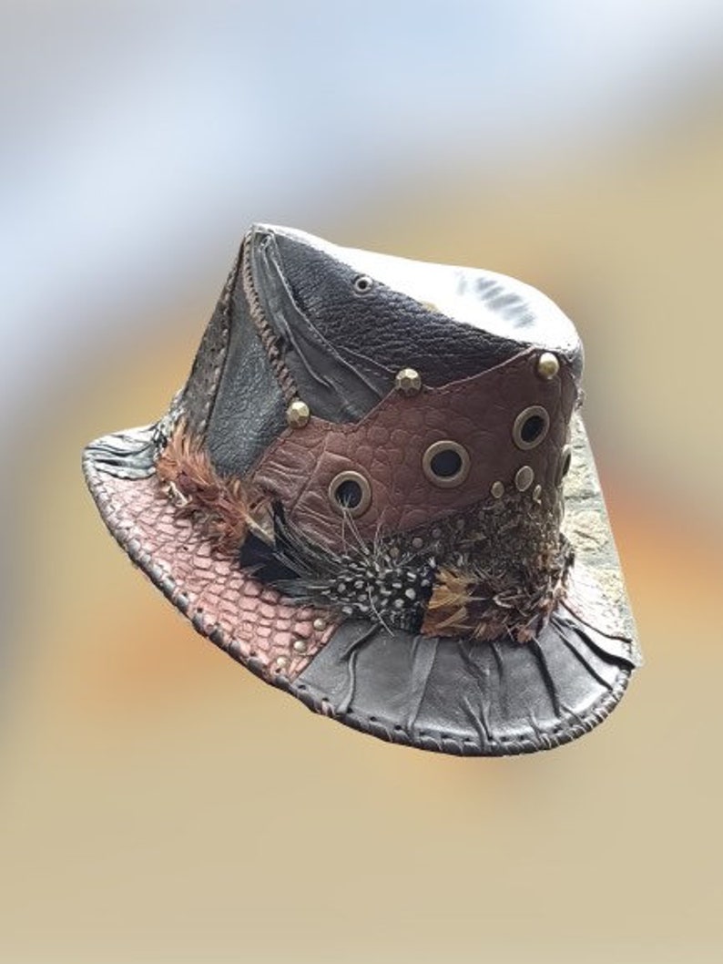 May include: A brown leather hat with a wide brim and a variety of textures and embellishments, including feathers, studs, and eyelets.