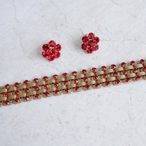 Könnte beinhalten: Ein goldenes Armband mit gewebtem Design und roten Strasssteinen. Zwei passende Ohrringe mit roten Strasssteinen sind ebenfalls enthalten.