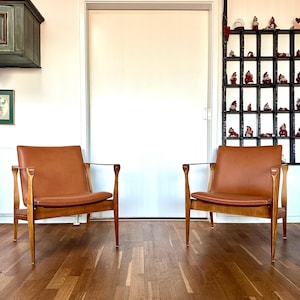May include: Two mid-century modern armchairs with brown leather seats and wooden frames. The chairs are positioned in front of a white door, with a wooden floor. A black shelf with small figurines is visible on the right.