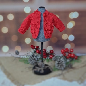 May include: A bright red knitted jacket with a round collar and button closure, displayed on a gray mannequin. The jacket features textured detailing on the front and sleeves. The background includes decorative elements like red berries and pinecones.