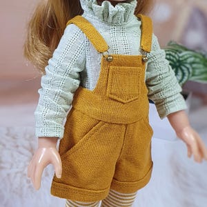 May include: A doll wearing a mustard yellow overall shorts set with a front pocket and metal buttons, paired with a light mint green long-sleeved shirt featuring a ruffled collar and textured fabric, and white and tan striped leggings.
