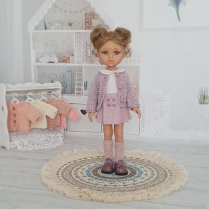May include: A doll dressed in a light purple knitted cardigan, matching skirt, and striped socks. The doll has brown hair styled in two buns and is wearing brown shoes. A miniature dollhouse and a rack of miniature sweaters are in the background.