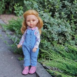 May include: A doll with shoulder-length reddish-blonde hair, brown eyes, and a light complexion. The doll wears a long-sleeved white shirt with a pink floral pattern under light blue corduroy overalls with silver buttons and pink lace-up shoes.
