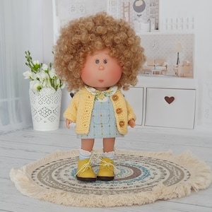 May include: A doll with curly blonde hair, wearing a light yellow knit cardigan with three buttons over a light blue patterned overall dress. The doll has a round face, blue eyes, and is wearing white socks with yellow lace-up shoes.