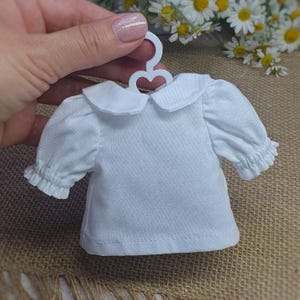 May include: A small white doll's blouse with a Peter Pan collar and frilled sleeves, suspended from a tiny white hanger. The blouse is crafted from a soft material and has a delicate, handmade look. The background includes daisies and a hessian surface.