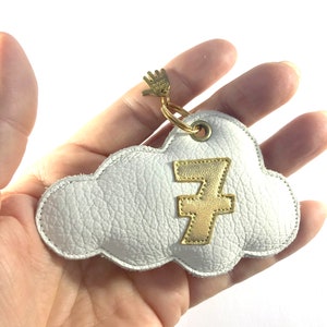 May include: White cloud-shaped keyring with a gold number 7 applique. The keyring is made of leather and has a gold-coloured metal ring and charm. The charm is a small hand. The keyring is held in a hand.
