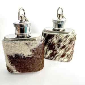 Op de afbeelding: Twee roestvrijstalen heupflacons met een koeienhuidleren wikkel. De flacons hebben een zilveren dop met een lus. De koeienhuid heeft een bruin-wit vlekkenpatroon.