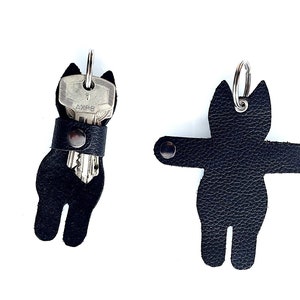 May include: Two black leather cat-shaped keyrings. One holds keys, secured by a snap. The other is a cat silhouette with a metal ring. Both keyrings have a textured surface and are designed to hold keys.