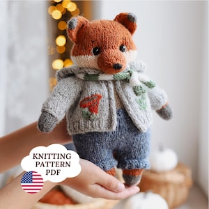 Może przedstawiać: Ręcznie robiona dziergana lalka lisa w szarej kurtce z czerwoną kwiatem, niebieskich spodniach i szaliku. Lis ma pomarańczowo-białe futro, czarne oczy i czarny nos. Obraz zawiera tekst "KNITTING PATTERN PDF" i amerykańską flagę.