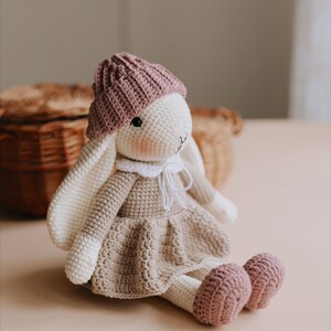 May include: A crocheted bunny wearing a pink hat and a brown dress. The bunny is sitting on a light brown surface.
