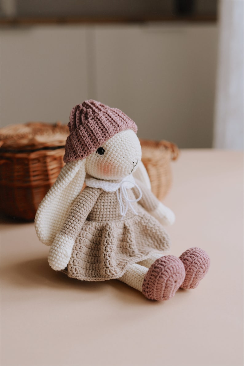 May include: A crocheted bunny wearing a pink hat and a brown dress. The bunny is sitting on a light brown surface.