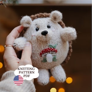 May include: A cream and brown knitted hedgehog toy with a mushroom applique. The hedgehog has black eyes, a black nose, and pink cheeks. The image includes a speech bubble with the text "KNITTING PATTERN PDF" and an American flag.