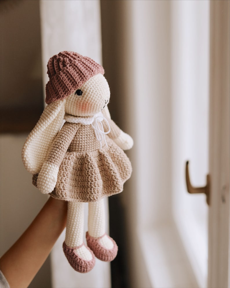 Puede incluir: Una mu&ntilde;eca de conejo de crochet con un sombrero rosa y un vestido marr&oacute;n. La mu&ntilde;eca tiene pelo blanco y mejillas rosadas.