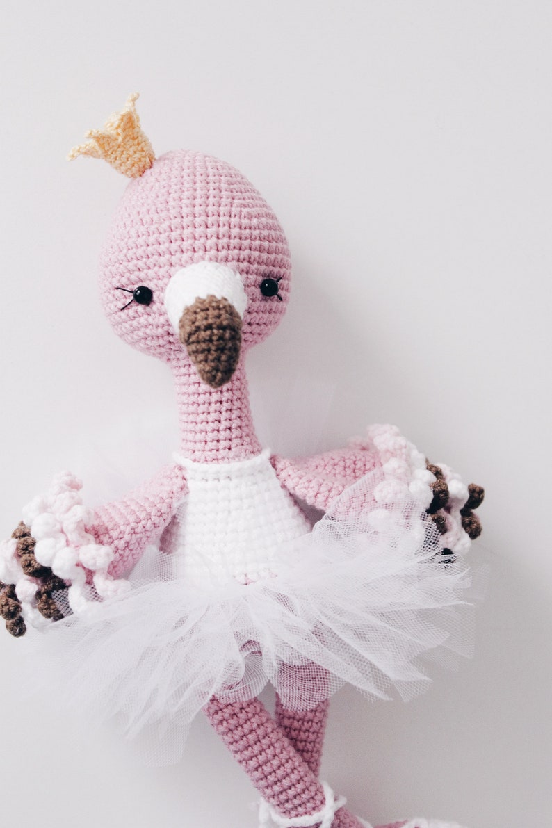 May include: A pink crocheted flamingo doll wearing a white tutu and a yellow crown. The doll has black eyes and a brown beak.