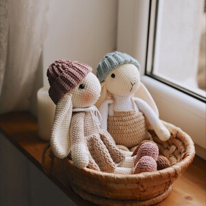 May include: Two crocheted bunny toys with pink and blue hats sitting in a wicker basket. The bunnies are wearing brown and white outfits.