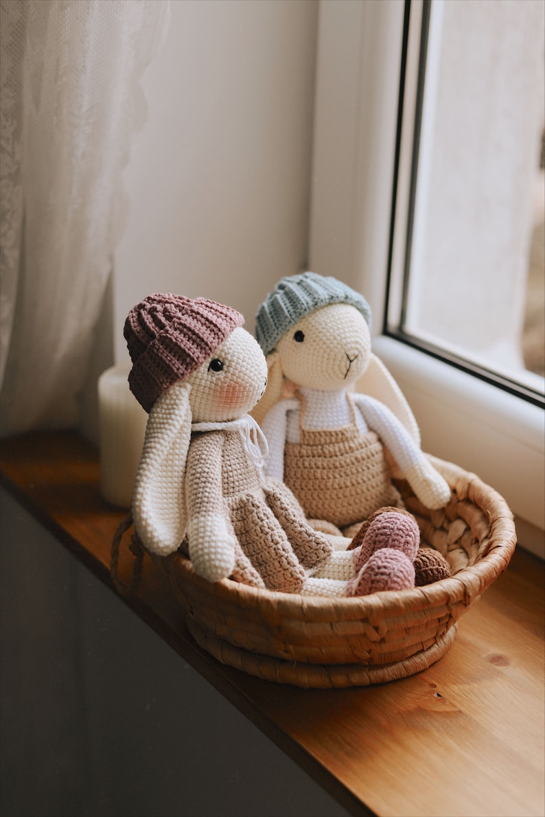 May include: Two crocheted bunny toys with pink and blue hats sitting in a wicker basket. The bunnies are wearing brown and white outfits.