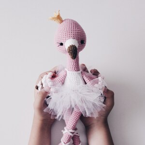 May include: A pink crocheted flamingo doll wearing a white tulle tutu and a gold crown. The doll is being held by two hands.
