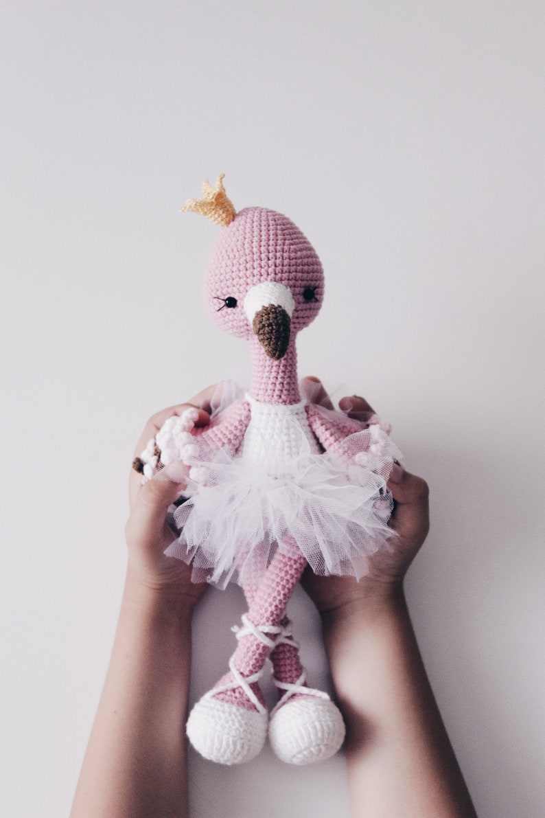 May include: A pink crocheted flamingo doll wearing a white tulle tutu and a gold crown. The doll is being held by two hands.