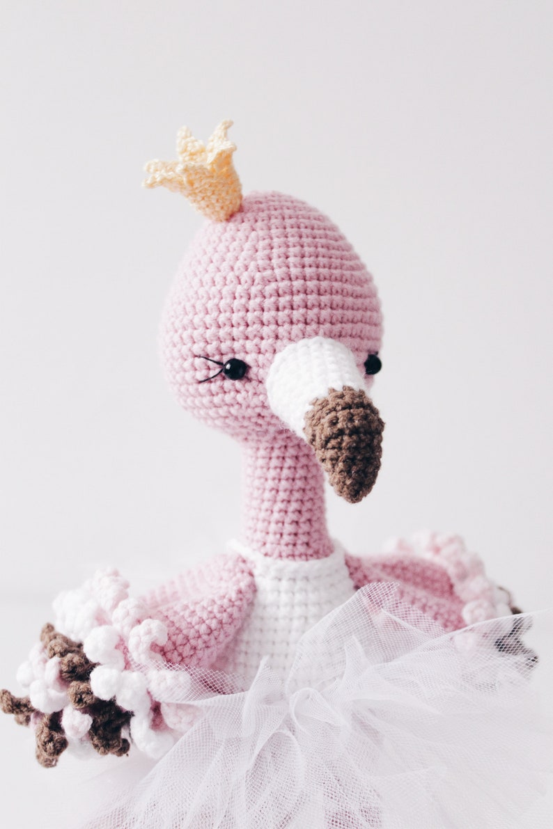 May include: A pink crocheted flamingo wearing a yellow crown and a white tulle tutu. The flamingo has black eyes and a brown beak.
