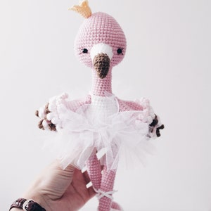 May include: Pink crocheted flamingo doll wearing a white tulle tutu and a gold crown. The flamingo has brown eyes and a brown beak. The doll is wearing white ballet shoes.
