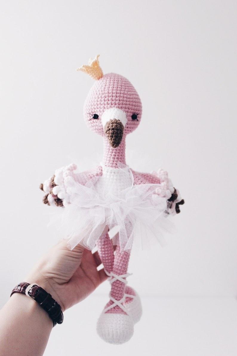 May include: Pink crocheted flamingo doll wearing a white tulle tutu and a gold crown. The flamingo has brown eyes and a brown beak. The doll is wearing white ballet shoes.
