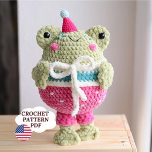 May include: A handmade crochet frog doll with a party hat. The frog is primarily light green with pink cheeks, a white and blue striped top, and pink pants with white polka dots. The doll has large black eyes and a white bow. The text "CROCHET PATTERN PDF" is in a speech bubble.