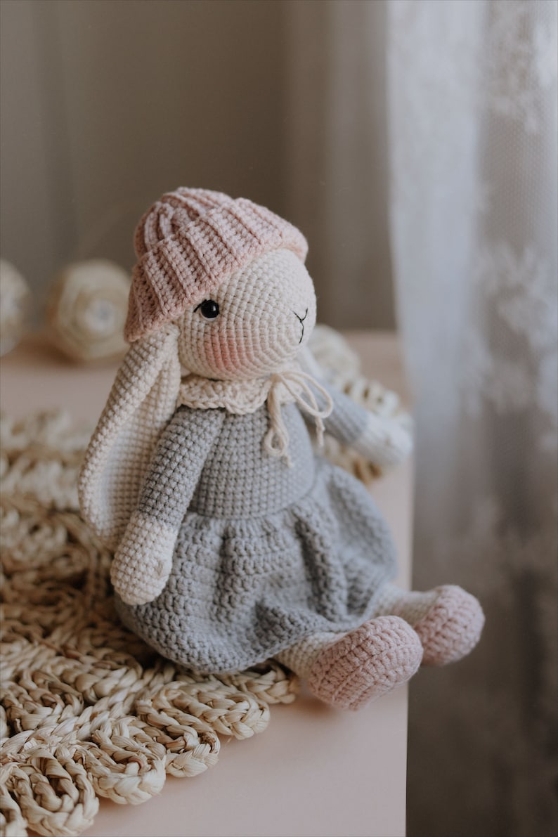 Puede incluir: Un juguete de conejo de crochet con un sombrero rosa y un vestido gris. El conejo est&aacute; sentado en una estera de paja tejida.