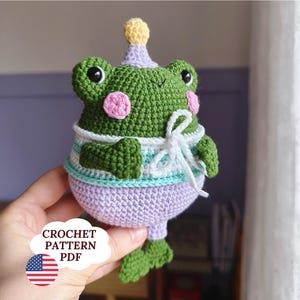 May include: A handmade green crochet frog wearing a party hat and a sweater with blue and white stripes. The frog has pink cheeks and is holding a white bow. The image includes the text "CROCHET PATTERN PDF" and an American flag.