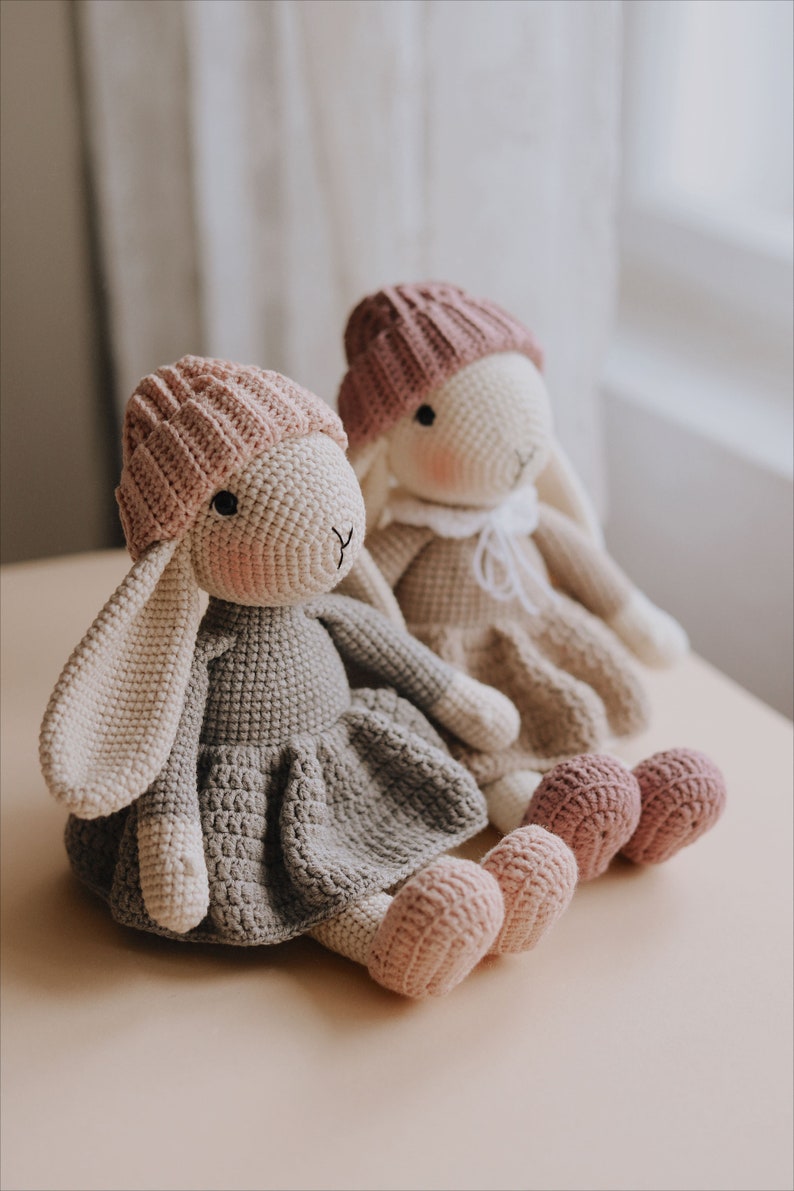 Puede incluir: Dos mu&ntilde;ecas de conejo de crochet con sombreros y vestidos rosas. El conejo de la izquierda lleva un vestido gris y el conejo de la derecha lleva un vestido blanco.