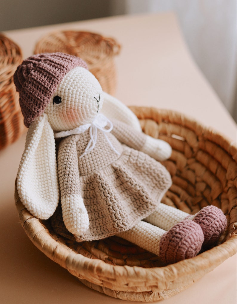 Puede incluir: Un juguete de conejo de crochet con un sombrero rosa y un vestido marr&oacute;n. El conejo est&aacute; sentado en una cesta tejida.