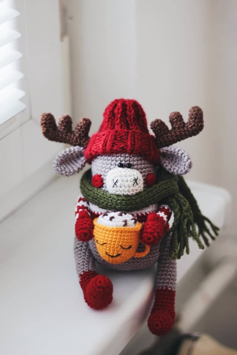 May include: A gray crocheted reindeer wearing a red hat and green scarf, holding a yellow mug with a smiling face and brown chocolate inside.