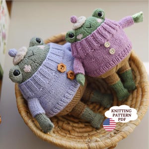 Op de afbeelding: Twee gebreide kikkerpoppen met zwarte ogen en roze wangen, gekleed in paarse truien met twee knopen, lichtbruine broeken, groene laarzen en kleine mutsjes. Een wolkvormige afbeelding met de tekst KNITTING PATTERN PDF is zichtbaar.