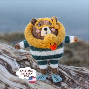 May include: A knitted bear toy wearing a yellow swim ring, yellow goggles, and a striped green and white swimsuit. The bear holds a small yellow duck toy with an orange beak. A white cloud-shaped label reads KNITTING PATTERN PDF with a US flag icon.