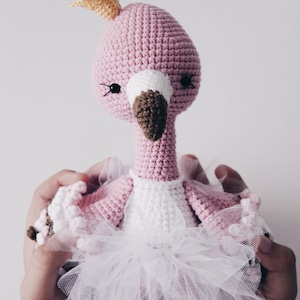 May include: A pink crocheted flamingo doll wearing a white tutu and a yellow crown. The doll is being held by two hands.