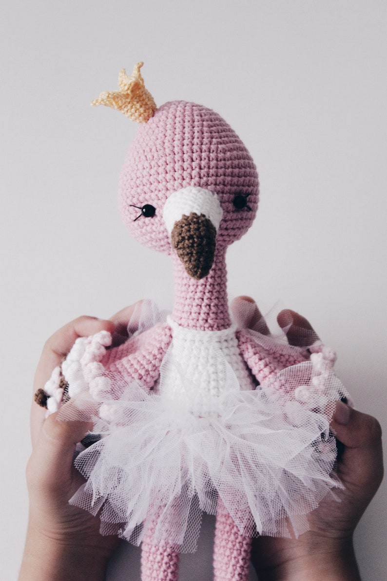 May include: A pink crocheted flamingo doll wearing a white tutu and a yellow crown. The doll is being held by two hands.