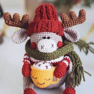 May include: A crocheted reindeer wearing a red hat, green scarf, and holding a yellow mug with a brown drink inside.