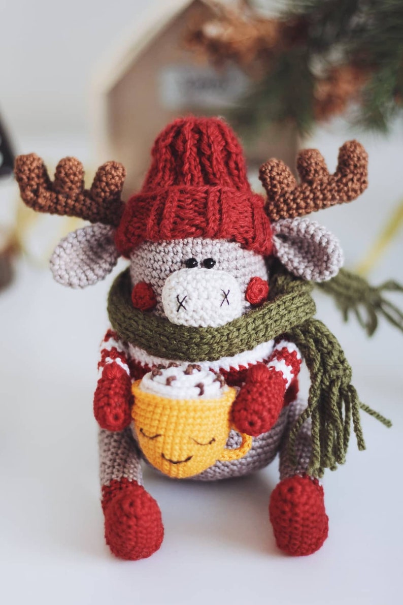 May include: A crocheted reindeer wearing a red hat, green scarf, and holding a yellow mug with a brown drink inside.