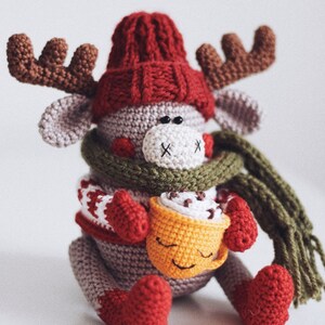 May include: A crocheted reindeer wearing a red hat, green scarf, and red mittens, holding a yellow crocheted mug with a brown swirl on top.