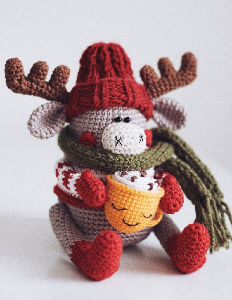 May include: A crocheted reindeer wearing a red hat, green scarf, and red mittens, holding a yellow crocheted mug with a brown swirl on top.