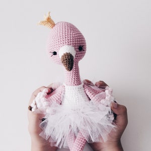 May include: A pink crocheted flamingo doll wearing a white tutu and a gold crown. The doll is being held by two hands.