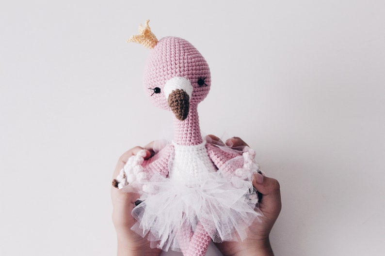 May include: A pink crocheted flamingo doll wearing a white tutu and a gold crown. The doll is being held by two hands.