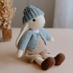 May include: A crocheted bunny toy wearing a blue knitted hat, a blue cardigan, and brown overalls. The bunny is sitting on a light brown surface.