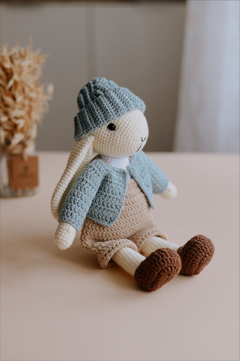 May include: A crocheted bunny toy wearing a blue knitted hat, a blue cardigan, and brown overalls. The bunny is sitting on a light brown surface.