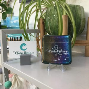 May include: A small, square, iridescent glass dish with a white outline of a person swimming in water. The dish is on a clear stand and has a "Claire Harris" business card in front of it.