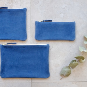 Könnte beinhalten: Drei blaue Wildleder-Reißverschlusstaschen in verschiedenen Größen. Die Taschen befinden sich auf einer hellgrauen Fliesenfläche mit einem Zweig Eukalyptusblätter.
