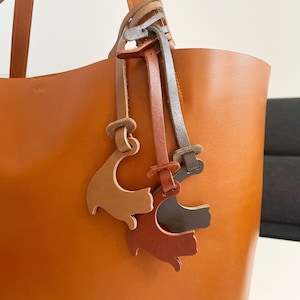 Peut inclure: Un sac cabas en cuir marron avec trois breloques en cuir en forme de chat attach&eacute;es &agrave; la poign&eacute;e. Les breloques sont en marron, orange et gris.