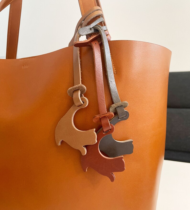 Peut inclure: Un sac cabas en cuir marron avec trois breloques en cuir en forme de chat attach&eacute;es &agrave; la poign&eacute;e. Les breloques sont en marron, orange et gris.